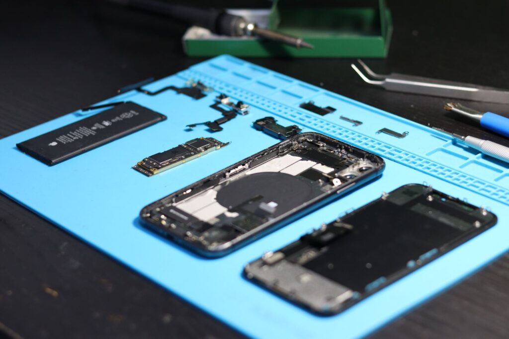 A smartphone taken apart on a blue repair mat with its battery, boards and screen laid out next to repair tools