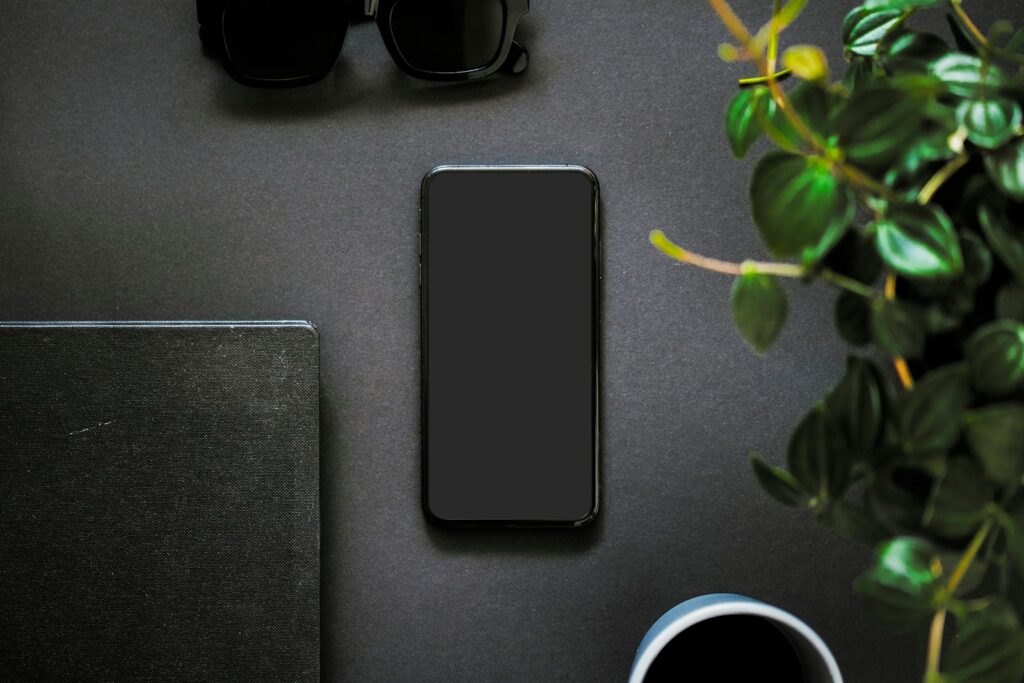 A black smartphone lying face up on a dark desk next to a notebook, sunglasses, a coffee cup and a green plant